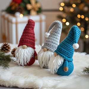 May include: Three handmade crochet gnomes in red, gray, and teal. Each gnome has a long, white beard, a small tan nose, and a matching hat with a white pom-pom. The gnomes are displayed on a white, fluffy surface, with a blurred background of Christmas decorations.