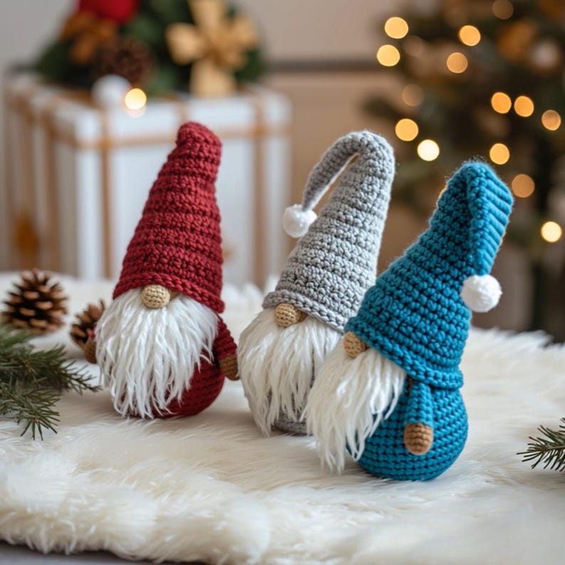 May include: Three handmade crochet gnomes in red, gray, and teal. Each gnome has a long, white beard, a small tan nose, and a matching hat with a white pom-pom. The gnomes are displayed on a white, fluffy surface, with a blurred background of Christmas decorations.