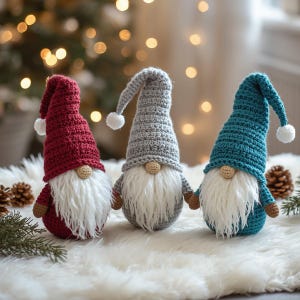 May include: Three handmade crochet gnomes in red, gray, and teal. Each gnome has a long, white beard, a small, tan nose, and a pointed hat with a white pom-pom. They are displayed on a white, fluffy surface.