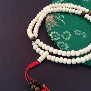 May include: A string of prayer beads featuring numerous smooth, off-white spherical beads, accented by several brown tiger eye gemstone beads. The strand is finished with a bright red knotted cord and two additional brown tiger eye beads at the end.