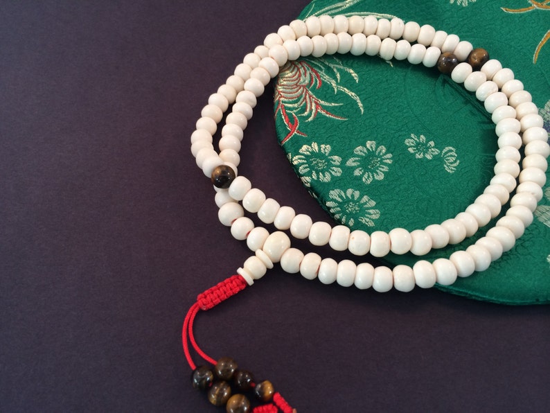 May include: A string of prayer beads featuring numerous smooth, off-white spherical beads, accented by several brown tiger eye gemstone beads. The strand is finished with a bright red knotted cord and two additional brown tiger eye beads at the end.