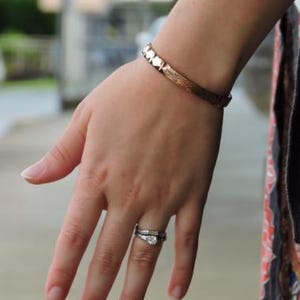 May include: A copper-colored bracelet with a geometric design is worn on a person's wrist. A silver ring with a round, clear stone is on the ring finger. The background is blurred, suggesting an outdoor setting.