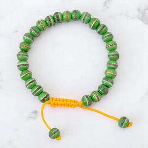 May include: Green beaded bracelet with gold accents and an adjustable yellow cord. The bracelet features round, green beads with white and gold detailing. The adjustable cord allows for a comfortable fit.