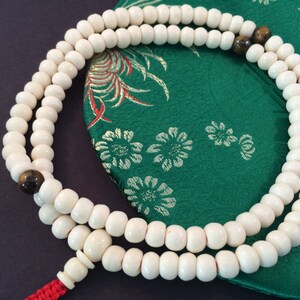 May include: A circular strand of off-white beads featuring two dark brown, polished accent beads. The strand is finished with a larger off-white bead and a bright red cord tassel, creating a continuous loop of prayer beads arranged in a simple pattern.