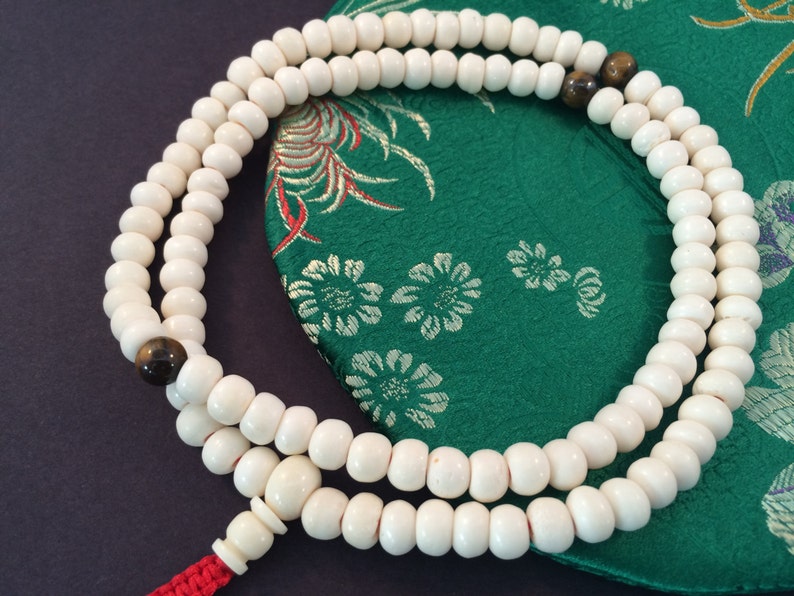 May include: A circular strand of off-white beads featuring two dark brown, polished accent beads. The strand is finished with a larger off-white bead and a bright red cord tassel, creating a continuous loop of prayer beads arranged in a simple pattern.