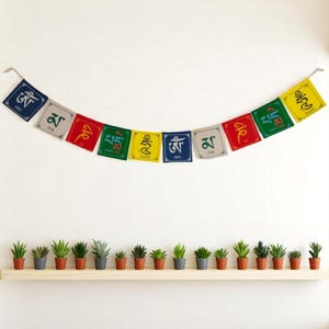 May include: A colorful string of prayer flags with various symbols and text hangs above a shelf of small potted succulents. The flags are in shades of blue, red, green, yellow, and white. The succulents are in small terracotta and gray pots.