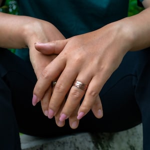 May include: A silver ring with a detailed design and a smaller gold ring are visible on a person's hands. The person is wearing a dark green top and black pants. The nails are painted with a light pink color.