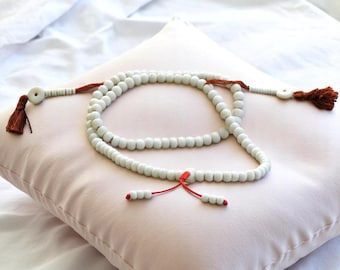 May include: A white beaded prayer rosary with brown tassels and a red accent, resting on a light pink pillow. The beads are evenly spaced, and the tassels are a deep brown color.