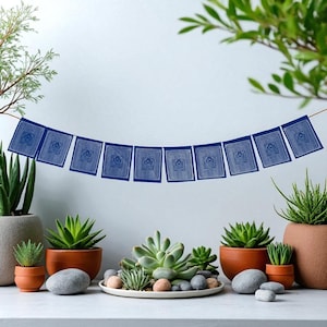 May include: A string of blue prayer flags with white intricate designs hangs above a display of potted succulents and decorative stones. The flags are square and evenly spaced, creating a serene and decorative scene.