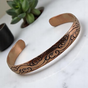 May include: Copper cuff bracelet with an engraved wave pattern. The open-ended bracelet has a smooth, polished finish and a decorative design along the top. The jewelry is displayed on a white marble surface.