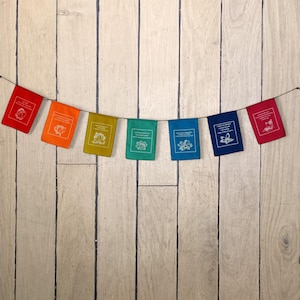 May include: A colorful banner of small square flags in red, orange, yellow-green, teal, blue, navy, and red. Each flag has a white floral design and text. The flags are strung on a black string against a light wood background.