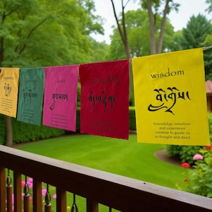 May include: A string of colorful prayer flags with words like "happiness", "love", and "wisdom" printed on them. The flags are strung across a wooden porch railing with a green lawn and trees in the background.