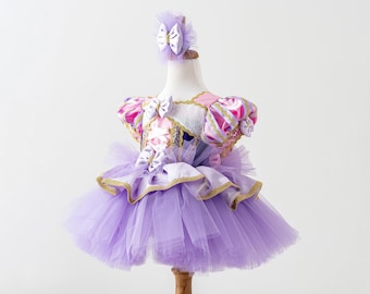 Puede incluir: Un vestido tutú lavanda y rosa con una diadema a juego, exhibido en un maniquí. El corpiño presenta detalles en rosa y dorado, con una falda de tul en capas. El vestido tiene un ribete dorado y está diseñado para una niña.