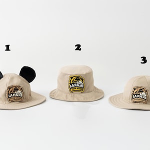 May include: Three beige safari hats with the text "SAFARI TOUR GUIDE" on a patch. One hat has black ears, the other two are standard bucket hats. The hats are on a white background.