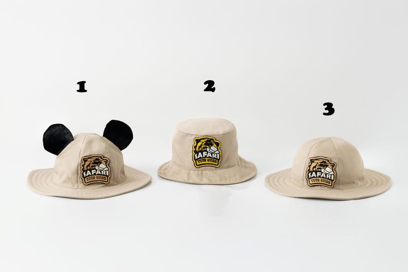 May include: Three beige safari hats with the text "SAFARI TOUR GUIDE" on a patch. One hat has black ears, the other two are standard bucket hats. The hats are on a white background.