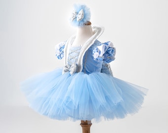 Puede incluir: Un vestido tutú azul claro con una diadema a juego, exhibido en un maniquí. El vestido presenta una falda de tul en capas, un corpiño adornado y mangas decorativas. La diadema tiene un lazo con detalles plateados. El conjunto es para una niña.