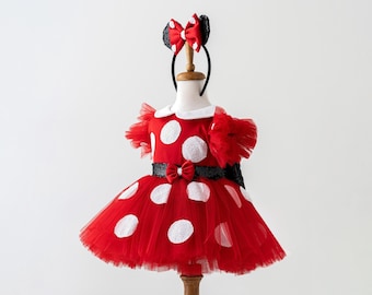 May include: A red polka dot dress with a tulle skirt and white collar, displayed on a mannequin. The dress features large white polka dots and a black sequined belt with a bow. A matching headband with bow-shaped ears completes the outfit.