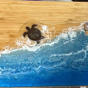 May include: A rectangular bamboo cutting board with a beach-themed design. The board features a brown turtle, seashells, and a blue and white resin ocean design. The resin simulates waves and sand.