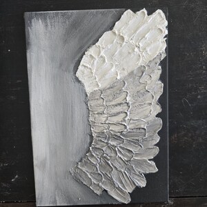 May include: A textured canvas art piece featuring a stylized angel wing. The wing is rendered in shades of white and gray, with a raised, layered texture. The background is a gradient of gray and black, creating a dramatic contrast.