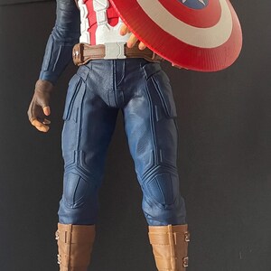 Puede incluir: Una estatua de Capit&aacute;n Am&eacute;rica en pose heroica, sosteniendo su ic&oacute;nico escudo. La figura viste un traje azul y blanco con botas y guantes marrones. La base dice "CAPTAIN AMERICA THE FIRST AVENGER."