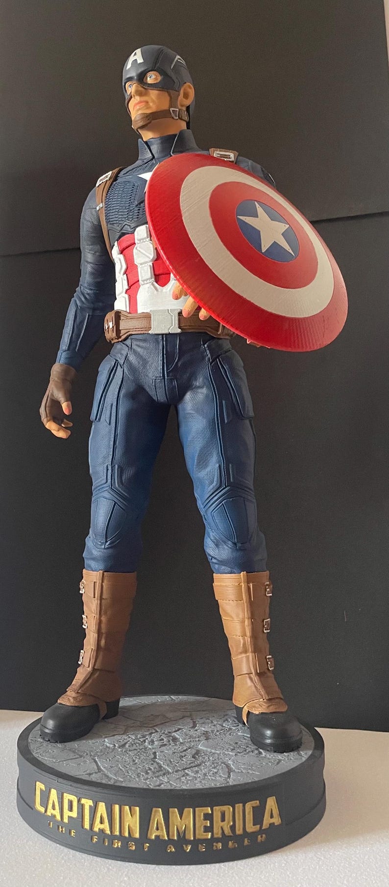 Puede incluir: Una estatua de Capit&aacute;n Am&eacute;rica en pose heroica, sosteniendo su ic&oacute;nico escudo. La figura viste un traje azul y blanco con botas y guantes marrones. La base dice "CAPTAIN AMERICA THE FIRST AVENGER."