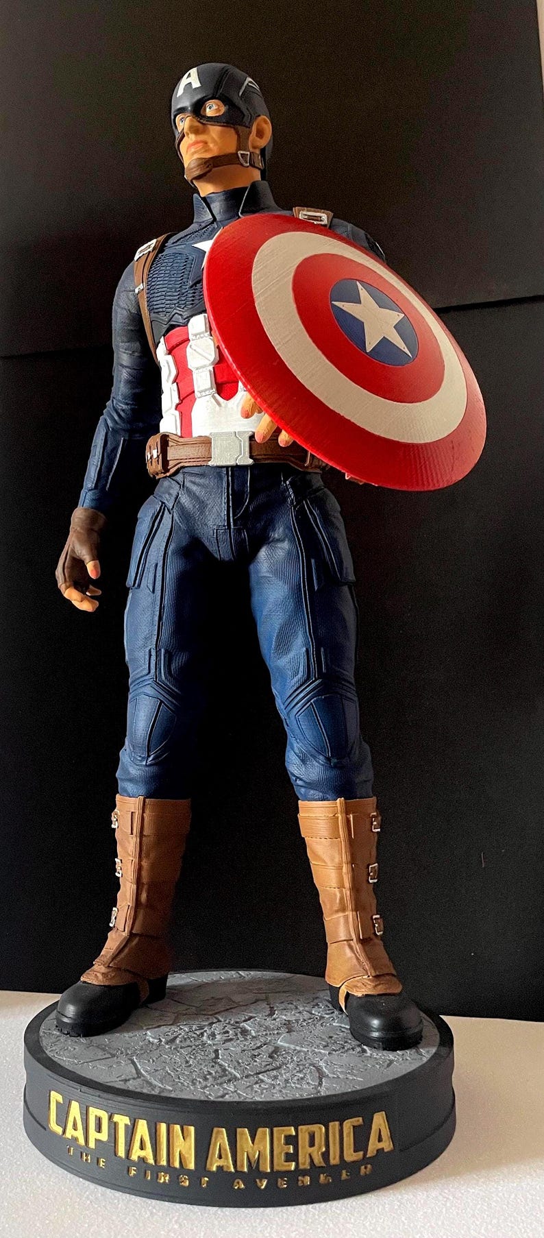 Puede incluir: Una figura de Capit&aacute;n Am&eacute;rica se encuentra sobre una base con el texto "CAPTAIN AMERICA THE FIRST AVENGER". La figura viste un uniforme azul y blanco, botas marrones y sostiene un escudo rojo, blanco y azul.
