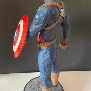 Puede incluir: Figura de Capit&aacute;n Am&eacute;rica, con un traje azul y rojo y un escudo. La figura est&aacute; sobre una base circular gris. El escudo es rojo con rayas blancas y una estrella azul. La figura mide aproximadamente 30 cm de altura.