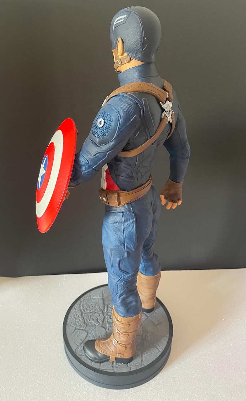 Puede incluir: Figura de Capit&aacute;n Am&eacute;rica, con un traje azul y rojo y un escudo. La figura est&aacute; sobre una base circular gris. El escudo es rojo con rayas blancas y una estrella azul. La figura mide aproximadamente 30 cm de altura.