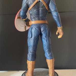 Puede incluir: Una figura de Capit&aacute;n Am&eacute;rica, vista desde atr&aacute;s, de pie sobre una base redonda. La figura viste un traje azul con detalles marrones, guantes y botas. El escudo es rojo, blanco y azul. La figura mide aproximadamente 30 cm de altura.