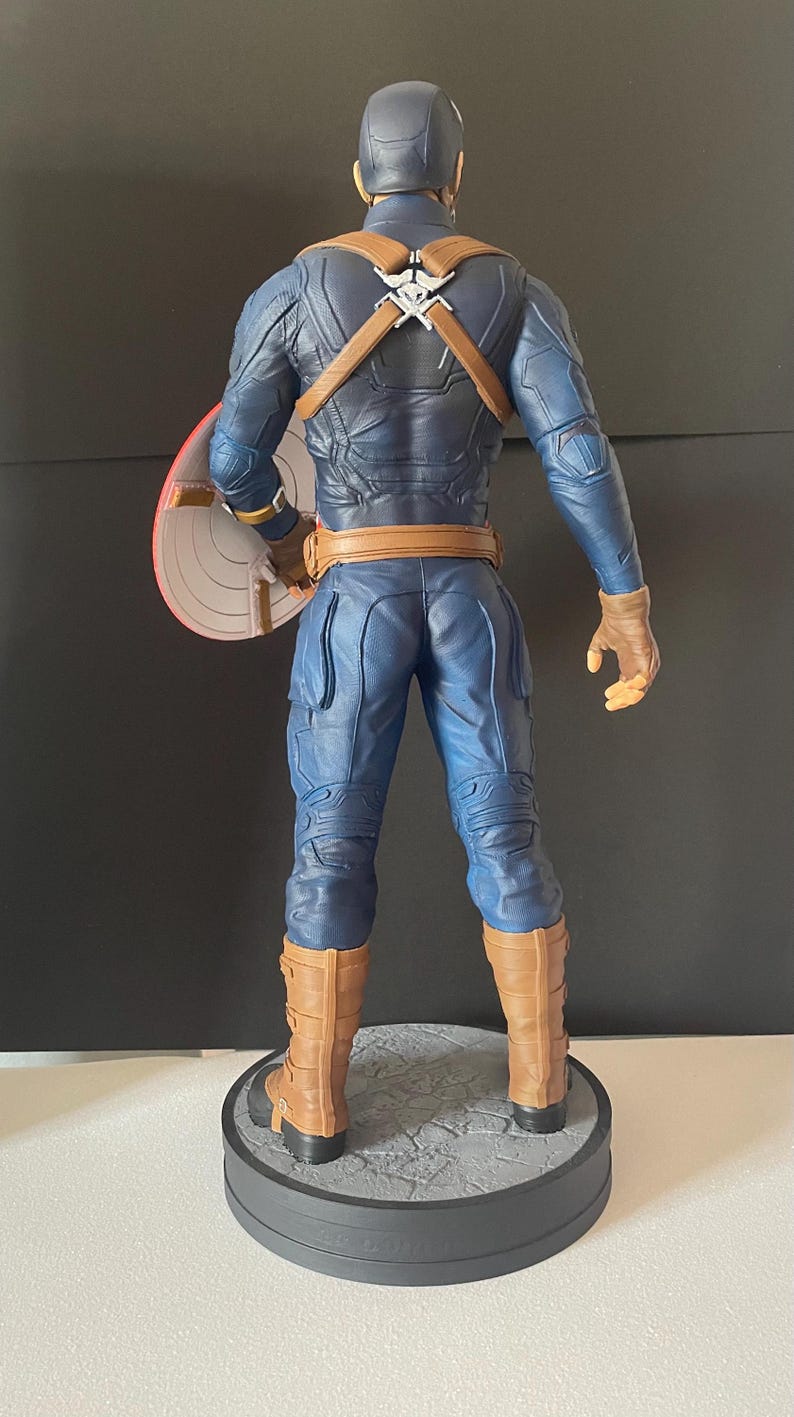 Puede incluir: Una figura de Capit&aacute;n Am&eacute;rica, vista desde atr&aacute;s, de pie sobre una base redonda. La figura viste un traje azul con detalles marrones, guantes y botas. El escudo es rojo, blanco y azul. La figura mide aproximadamente 30 cm de altura.