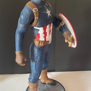 Puede incluir: Figura de Capit&aacute;n Am&eacute;rica en pose din&aacute;mica, con un traje azul y blanco con un emblema de estrella. La figura sostiene un escudo rojo, blanco y azul. La base es gris con la palabra "CAPTAIN". La figura mide aproximadamente 30 cm de altura.