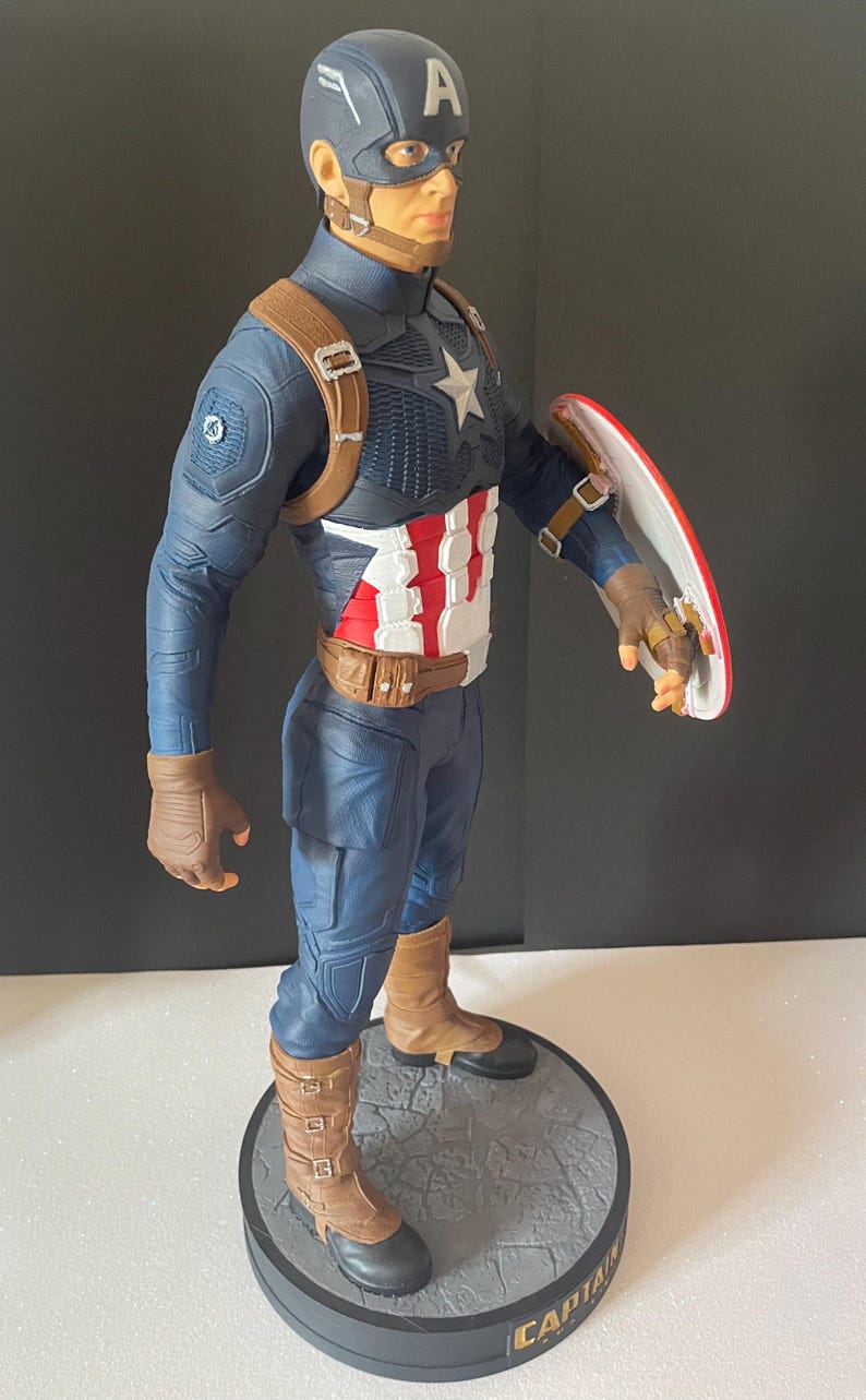 Puede incluir: Figura de Capit&aacute;n Am&eacute;rica en pose din&aacute;mica, con un traje azul y blanco con un emblema de estrella. La figura sostiene un escudo rojo, blanco y azul. La base es gris con la palabra "CAPTAIN". La figura mide aproximadamente 30 cm de altura.