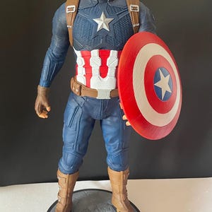 Puede incluir: Una figura de Capit&aacute;n Am&eacute;rica sobre una base redonda. La figura viste un traje azul y rojo con una estrella blanca en el pecho y un escudo a juego. La base dice "CAPTAIN AMERICA".