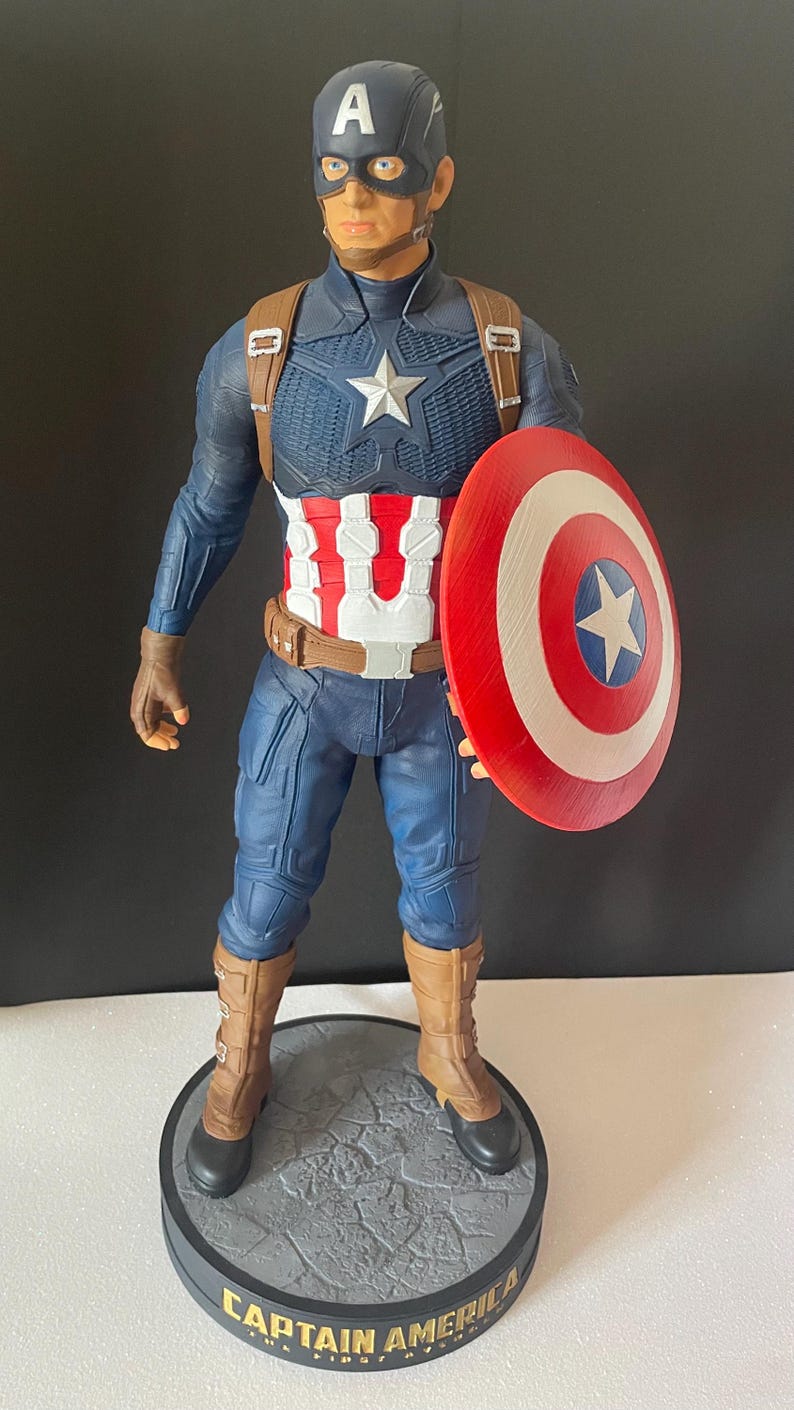 Puede incluir: Una figura de Capit&aacute;n Am&eacute;rica sobre una base redonda. La figura viste un traje azul y rojo con una estrella blanca en el pecho y un escudo a juego. La base dice "CAPTAIN AMERICA".