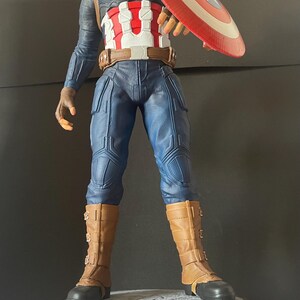 Puede incluir: Una figura de Capit&aacute;n Am&eacute;rica se encuentra sobre una base con letras doradas que dicen "CAPTAIN AMERICA THE FIRST AVENGER". La figura viste un uniforme azul y blanco, botas marrones y sostiene un escudo rojo, blanco y azul. La figura mide unos 30 cm de altura.
