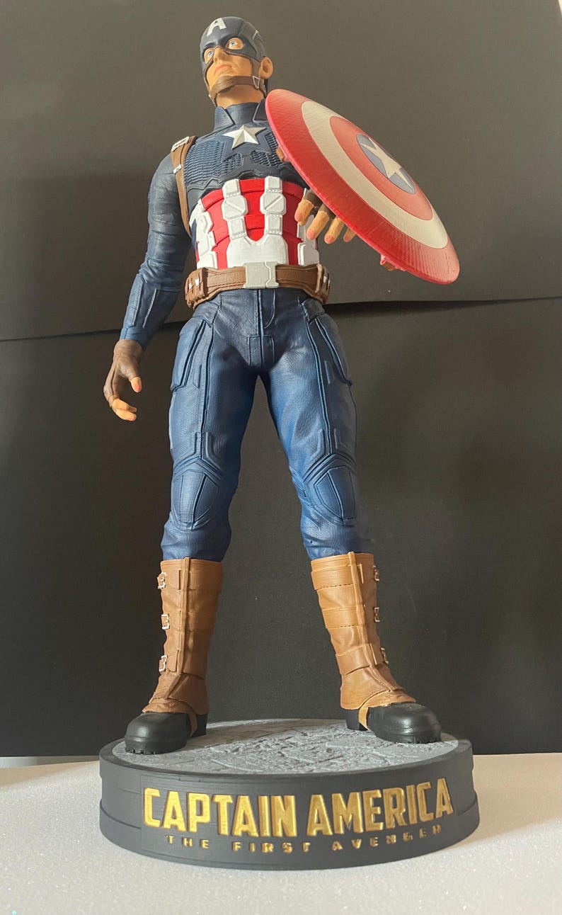 Puede incluir: Una figura de Capit&aacute;n Am&eacute;rica se encuentra sobre una base con letras doradas que dicen "CAPTAIN AMERICA THE FIRST AVENGER". La figura viste un uniforme azul y blanco, botas marrones y sostiene un escudo rojo, blanco y azul. La figura mide unos 30 cm de altura.