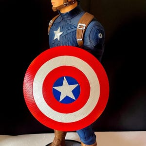 Puede incluir: Figura de acci&oacute;n de Capit&aacute;n Am&eacute;rica, con un traje azul y rojo, un escudo con una estrella y un casco con la letra "A". La figura est&aacute; posada sobre una base texturizada, mostrando detalles intrincados.