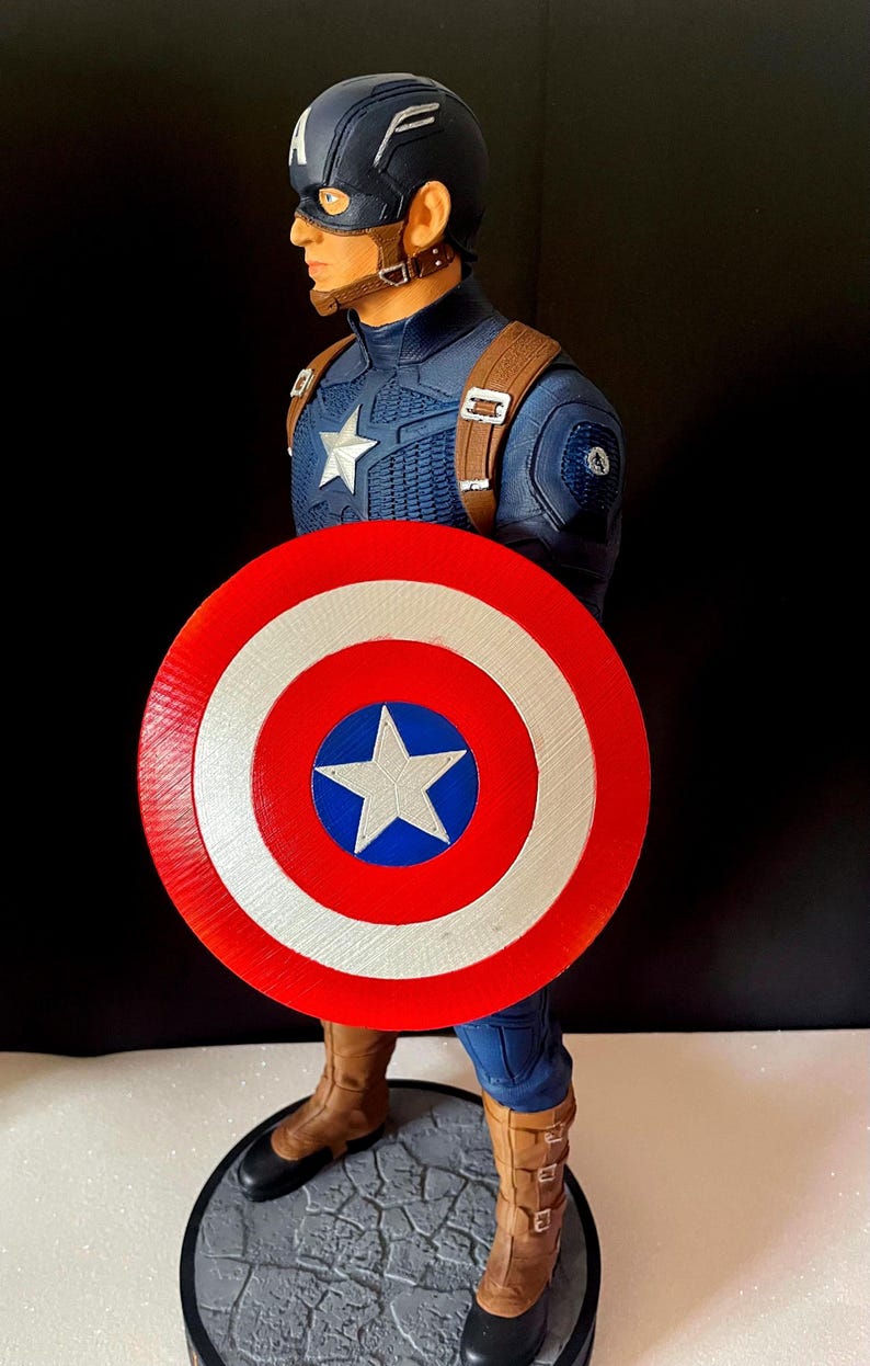 Puede incluir: Figura de acci&oacute;n de Capit&aacute;n Am&eacute;rica, con un traje azul y rojo, un escudo con una estrella y un casco con la letra "A". La figura est&aacute; posada sobre una base texturizada, mostrando detalles intrincados.