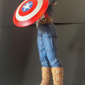 Puede incluir: Figura de Capit&aacute;n Am&eacute;rica, con un traje azul y rojo y un escudo. La figura est&aacute; sobre una base con la palabra "AMERICA". El escudo es rojo con una estrella blanca y un c&iacute;rculo azul. Las botas son marrones.