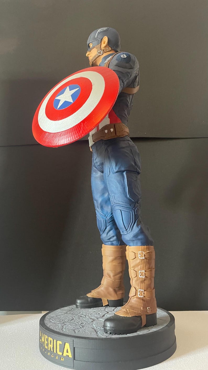 Puede incluir: Figura de Capit&aacute;n Am&eacute;rica, con un traje azul y rojo y un escudo. La figura est&aacute; sobre una base con la palabra "AMERICA". El escudo es rojo con una estrella blanca y un c&iacute;rculo azul. Las botas son marrones.