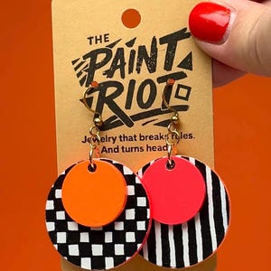 Può includere: Un paio di orecchini pendenti color oro con un motivo a scacchi bianchi e neri e un motivo a righe bianche e nere. Ogni orecchino ha un cerchio arancione brillante e un cerchio rosa brillante. Gli orecchini sono su una carta beige con il testo "THE PAINT RIOT".