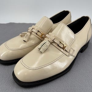 May include: A pair of cream-colored leather loafers with black soles. Each shoe features a decorative strap with a gold-tone buckle and a small tassel. The shoes have a glossy finish and are stitched with black thread.