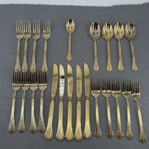 May include: A gold-toned flatware set consisting of eight forks, five knives, and five spoons. Each piece features a polished finish with a textured, vertical-lined pattern on the handle and a scalloped detail at the base of the handle.