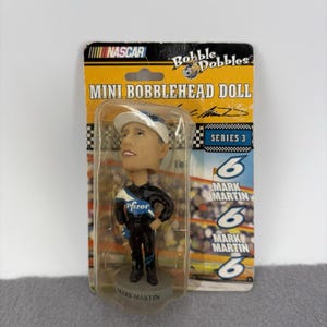 May include: A mini bobblehead doll of Mark Martin in a NASCAR racing suit, packaged in a clear plastic case. The doll has a white cap and the Pfizer logo on the front. The packaging includes the NASCAR and Bobble Dobbles logos.