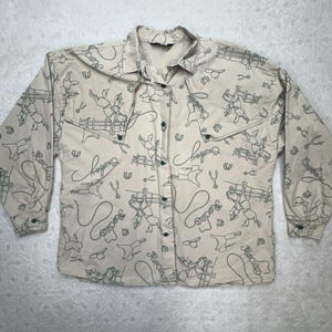 Pode incluir: Camisa bege com botões com um padrão de contorno verde repetido de cowboys, cavalos e equipamentos de rodeio. A palavra "Cowboy" está escrita em itálico. A camisa tem mangas compridas e botões verdes.