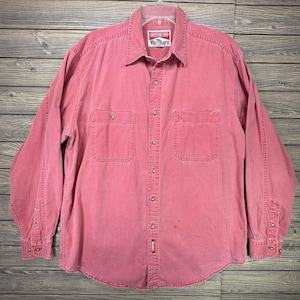 May include: A long-sleeved button-down shirt in a faded red colour. The shirt features a pointed collar, two chest pockets with button closures, and a front button placket. A label at the neck reads Adventure Team Marlboro, and a small tag is at the hem.