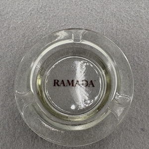 May include: A clear glass ashtray with the word "RAMADA" printed in red. The ashtray is circular with three notches for cigarettes. The ashtray is made of clear glass.