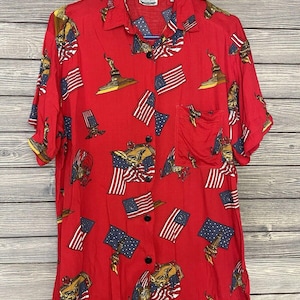 Puede incluir: Camisa roja con estampado patriótico. El patrón presenta imágenes repetidas de la bandera estadounidense, la Estatua de la Libertad y águilas. La camisa de manga corta tiene un bolsillo en el pecho y botones negros.