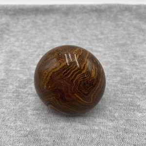 May include: A brown, rounded stone with a unique pattern of swirling, layered bands in shades of brown and tan. The stone has a smooth surface and a few white markings. The stone is a natural, earthy object.
