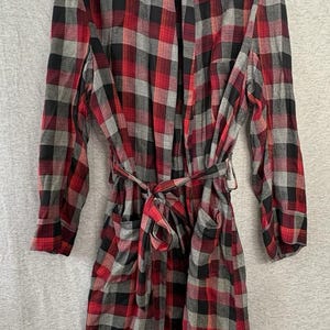 May include: A long-sleeved robe featuring a red, black, and gray plaid pattern. The garment includes a shawl collar, a matching fabric tie belt at the waist, and two front patch pockets. The robe is displayed on a hanger against a neutral background.